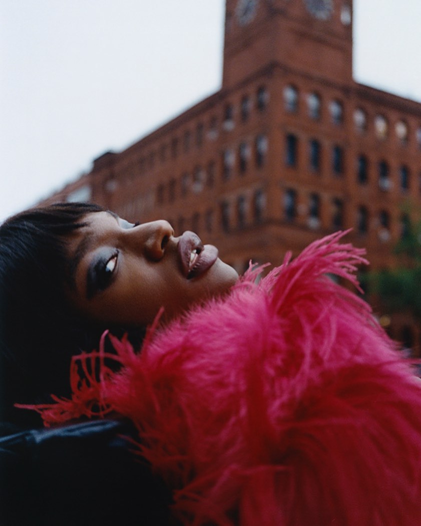 Portrait of a stylish Black woman with vibrant pink boa posing in front of a brick building with a clock tower on a cloudy day.