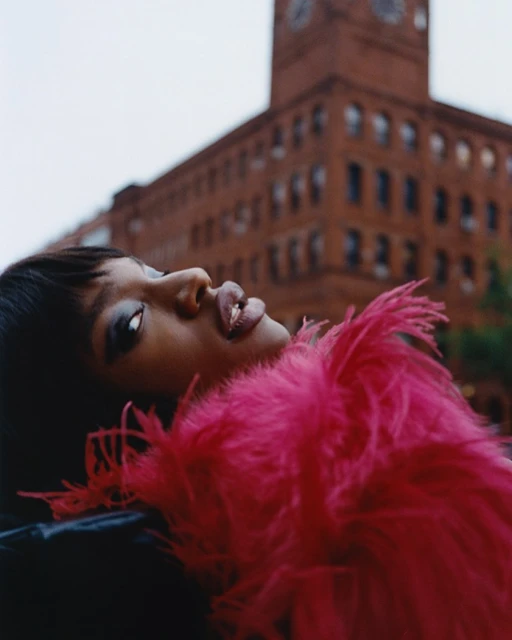 Portrait of a stylish Black woman with vibrant pink boa posing in front of a brick building with a clock tower on a cloudy day.