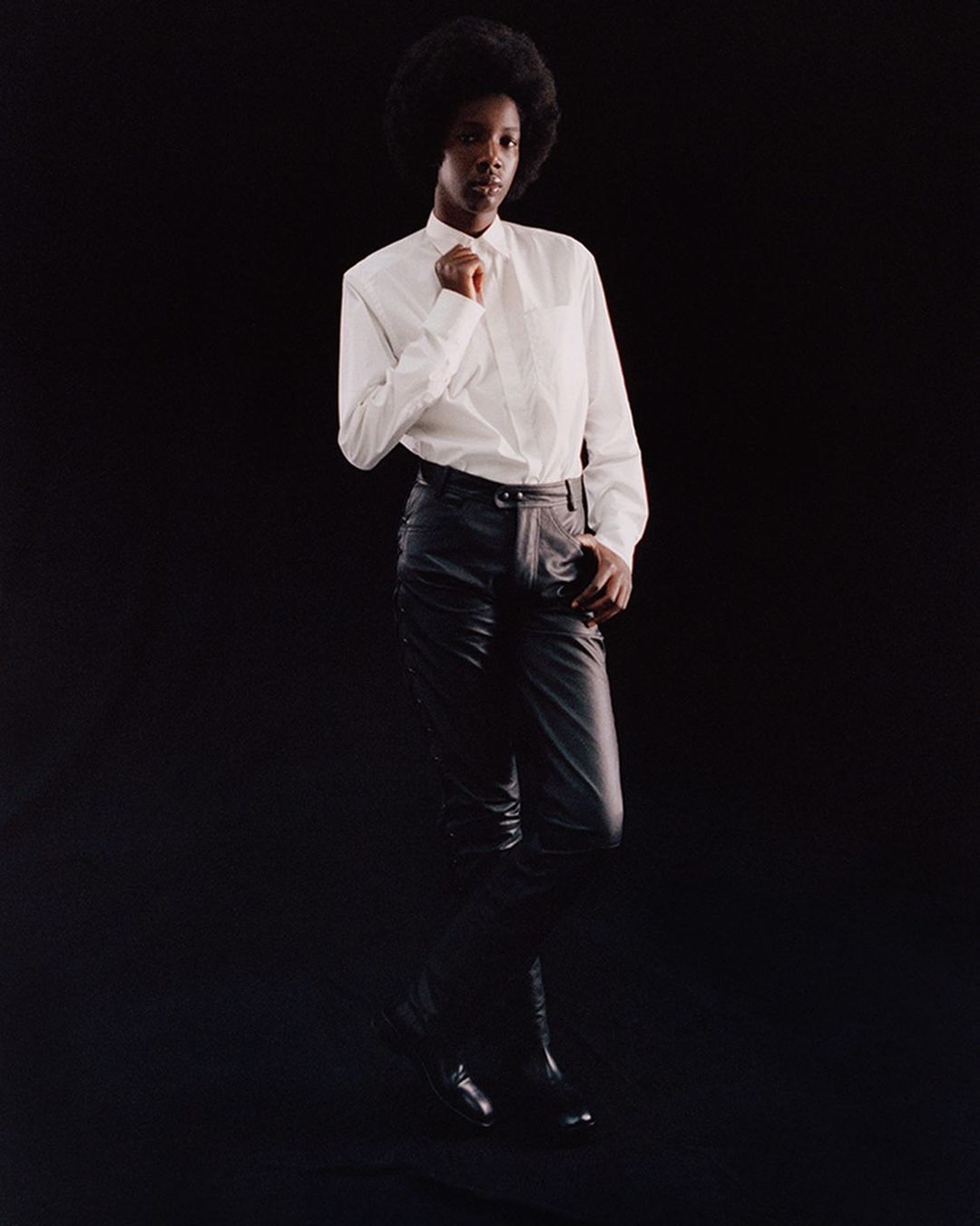 Model with afro wearing a white button-down shirt and black leather pants against a black background