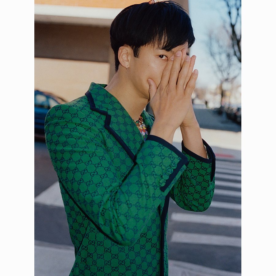 Adult Asian male in a green and blue Gucci patterned blazer poses outdoors, covering his face with his hands near a city street.