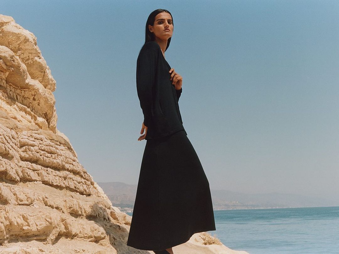 A slender woman in a long black dress stands gracefully on rocky terrain, against a backdrop of a clear blue sky and tranquil ocean.