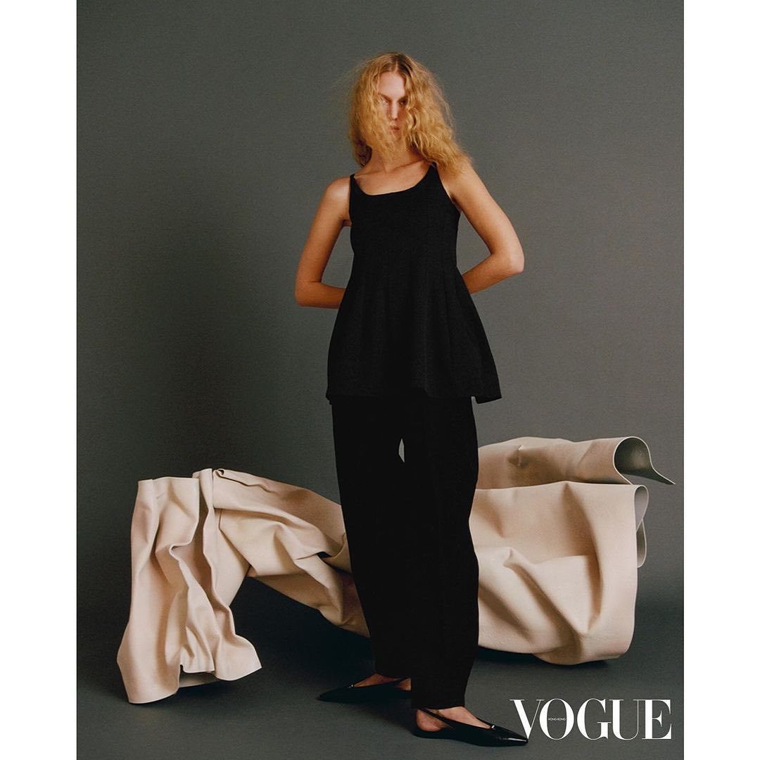 Fashion editorial shot of a blonde adult female model in black clothing, posing in front of draped beige fabric in a studio setting.