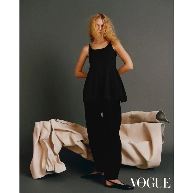 Fashion editorial shot of a blonde adult female model in black clothing, posing in front of draped beige fabric in a studio setting.