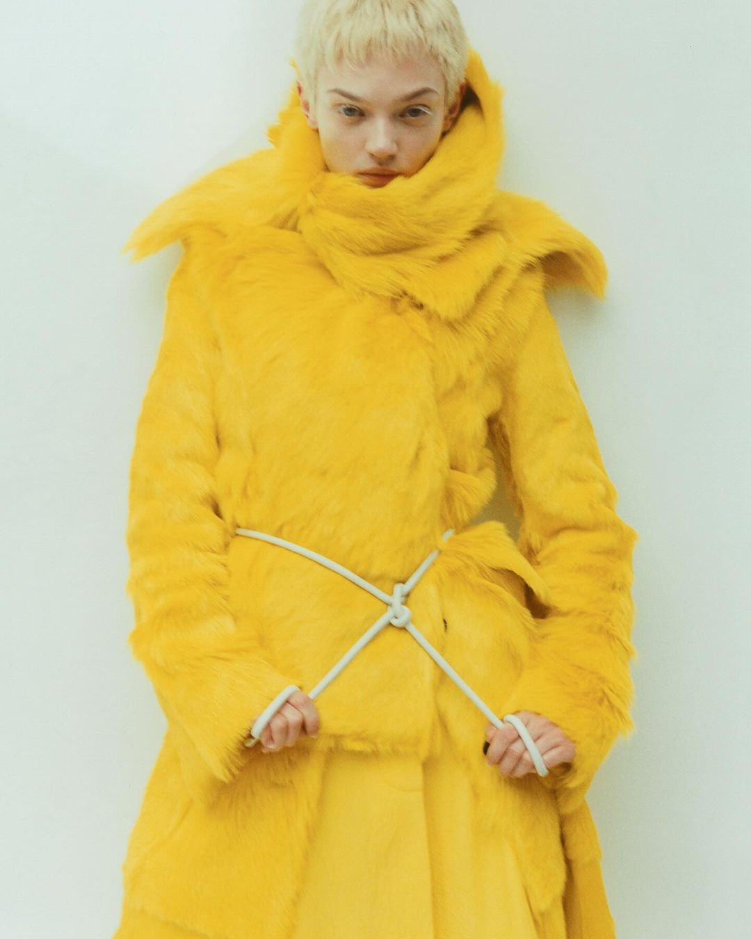 A fashion model poses in a bright yellow, fur-like coat with a white rope belt, against a white wall, in a studio shot.