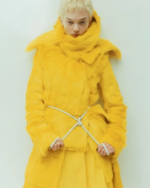 A fashion model poses in a bright yellow, fur-like coat with a white rope belt, against a white wall, in a studio shot.