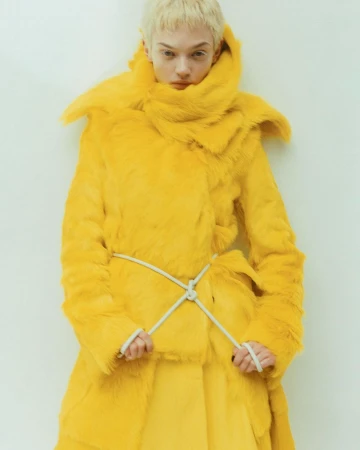 A fashion model poses in a bright yellow, fur-like coat with a white rope belt, against a white wall, in a studio shot.