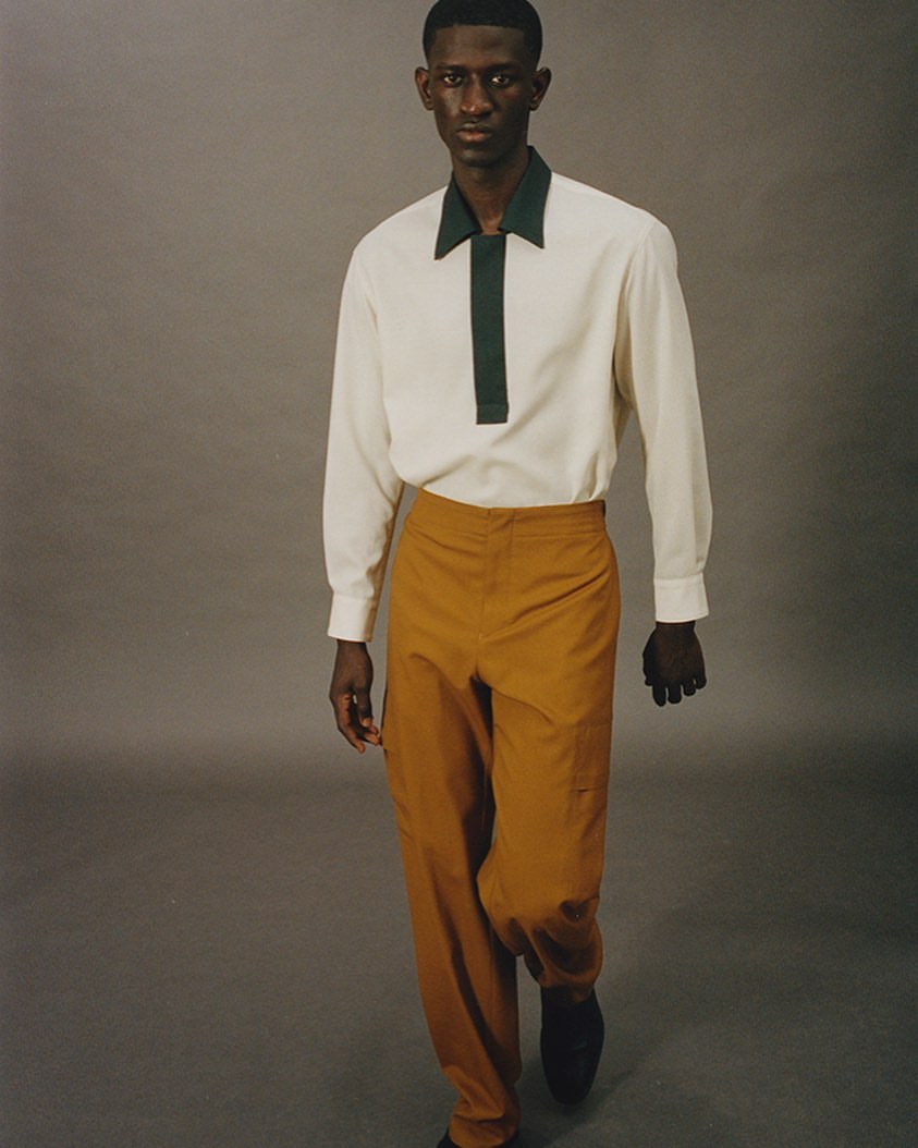 A dark-skinned man in a cream-colored shirt with green accents and tan pants is in a fashion studio, posing in a fashionable outfit.