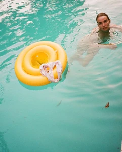 A woman in a turquoise swimming pool next to a bright yellow inflatable ring with clothes draped over it.