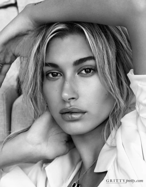 Black and white portrait of a model with hand on head, wearing a white shirt and necklace against a neutral, soft-lit studio backdrop.