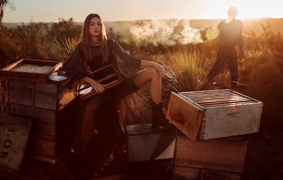 A woman poses on a wooden hive box near a beekeeper at sunset in a golden landscape, wearing a flowing dress and boots.