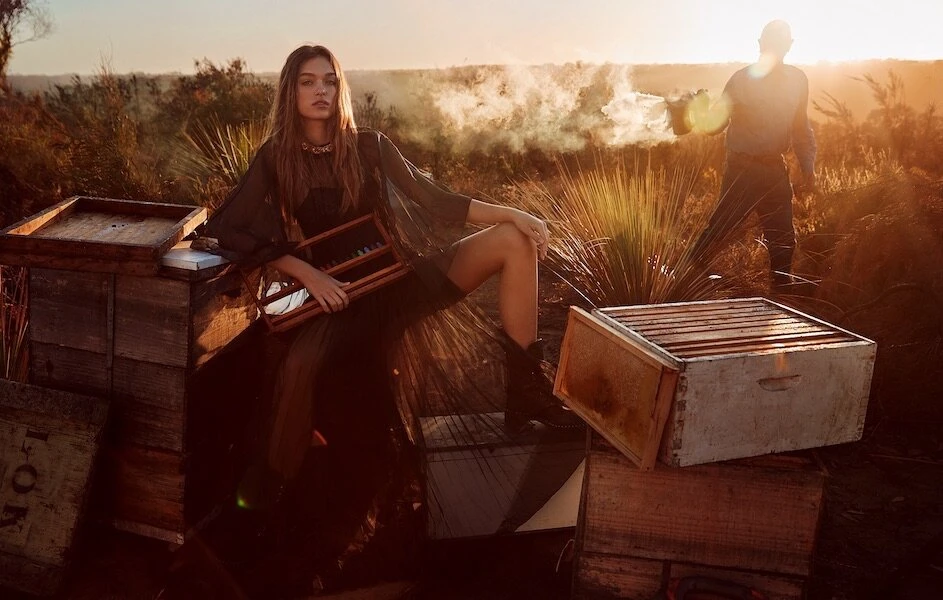 A woman poses on a wooden hive box near a beekeeper at sunset in a golden landscape, wearing a flowing dress and boots.