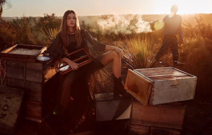 A woman poses on a wooden hive box near a beekeeper at sunset in a golden landscape, wearing a flowing dress and boots.