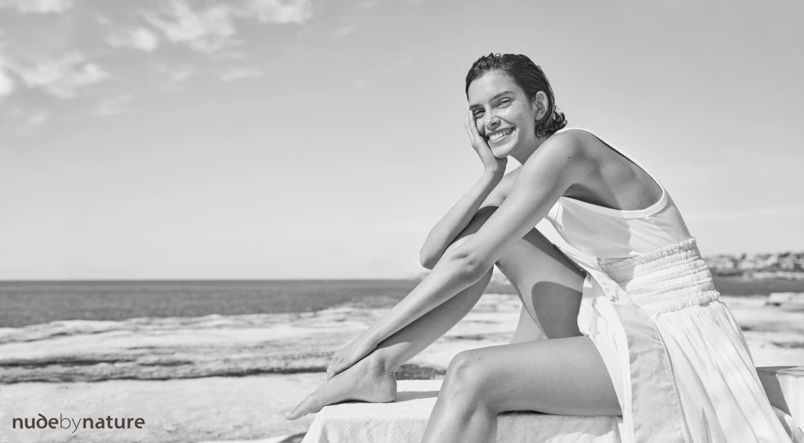 Monochrome portrait of a smiling woman in a white dress sitting outdoors by the ocean against a partly cloudy sky.