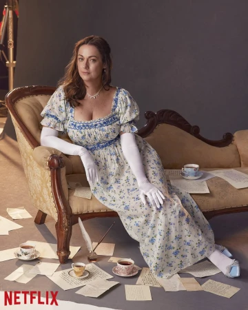 Woman in floral dress sitting on vintage chaise lounge with scattered papers and tea cups in studio setting. Netflix logo visible.