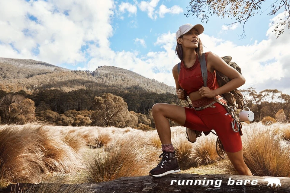Athletic woman in sportswear and baseball cap hiking through a grassy field with a mountain backdrop on a sunny day.