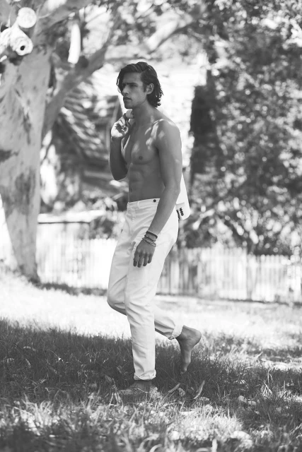Black and white portrait of a shirtless man walking in grass, wearing white pants, with a jacket slung over his shoulder outdoors.