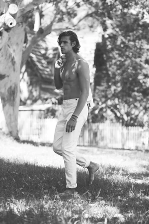 Black and white portrait of a shirtless man walking in grass, wearing white pants, with a jacket slung over his shoulder outdoors.