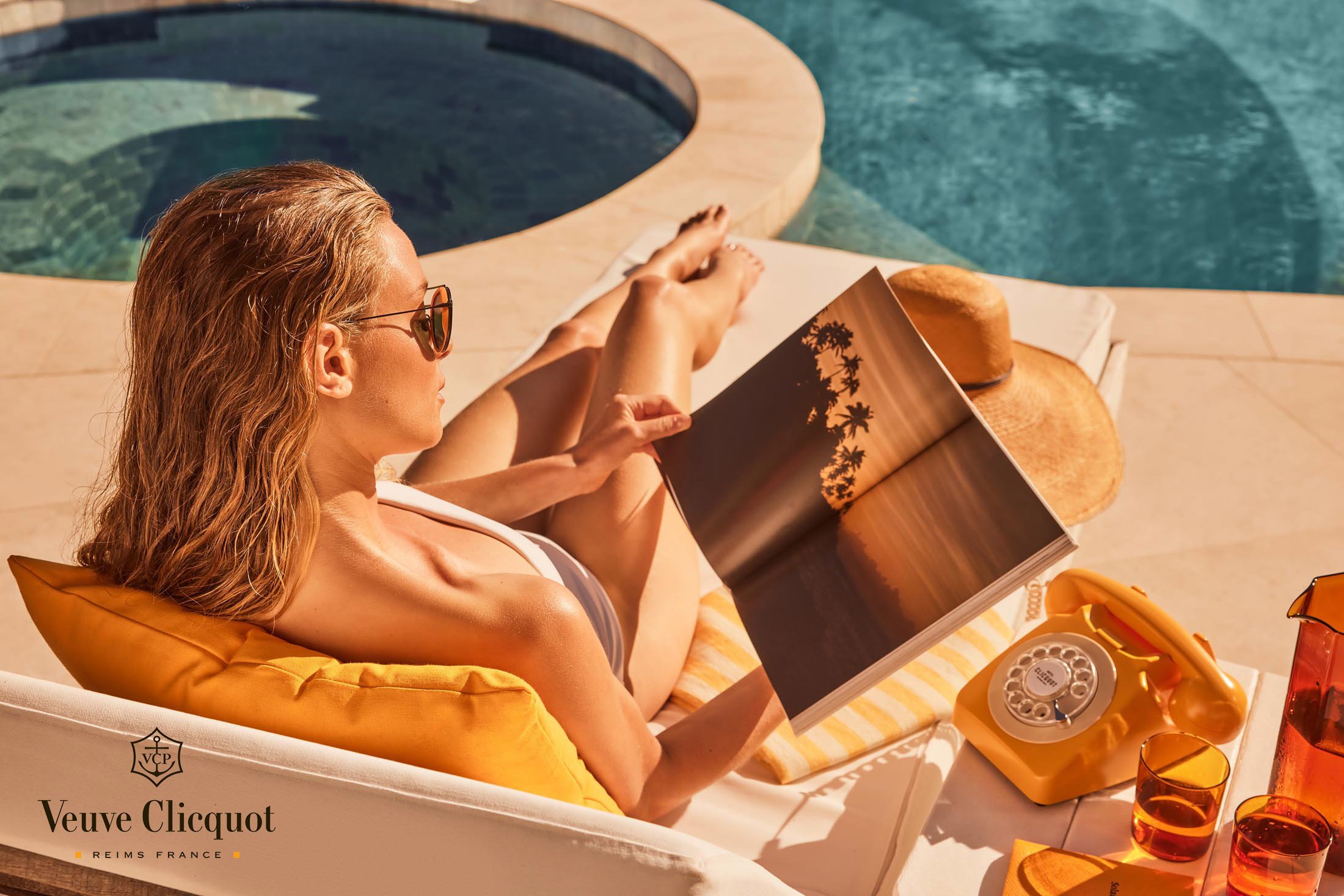 Woman in sunglasses lounges by the pool reading a magazine. Outdoor setting with bright sunlight, featuring poolside furniture.