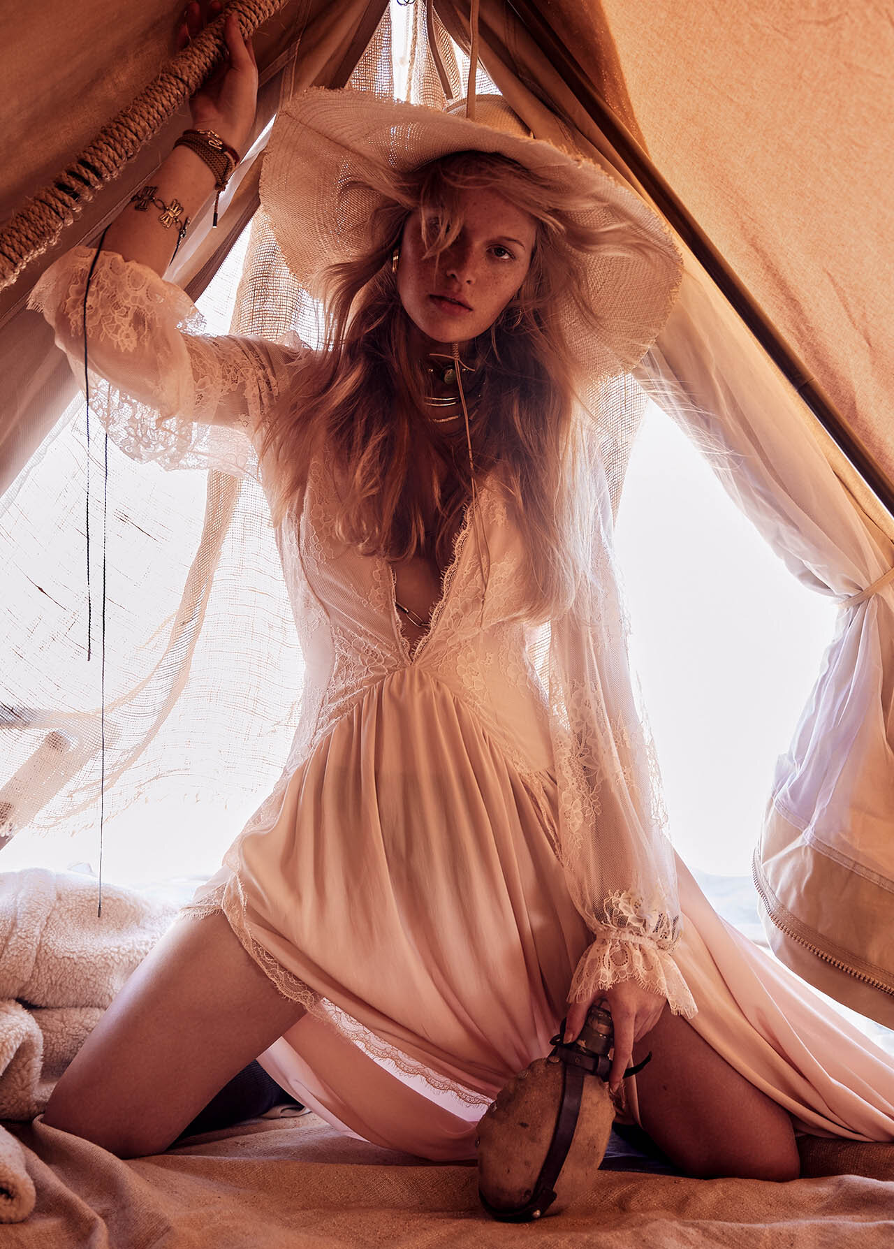 A blonde woman in a lace dress and straw hat poses inside a tent holding a water bottle, evoking a dreamy, bohemian fashion vibe.