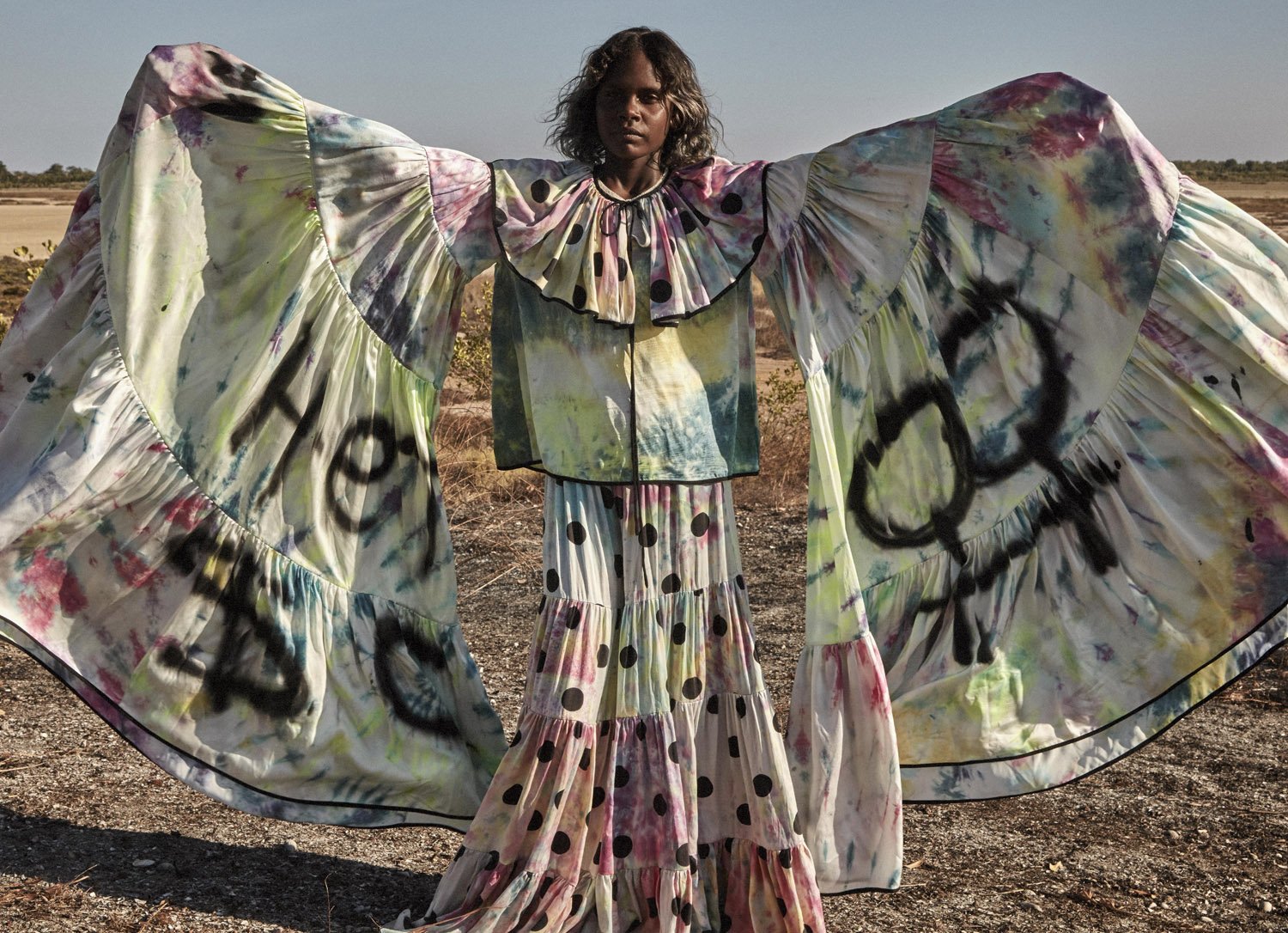 An indigenous woman in a full-length polka dot dress and tie dye print jacket with large sleeves stands in an open, rural landscape.