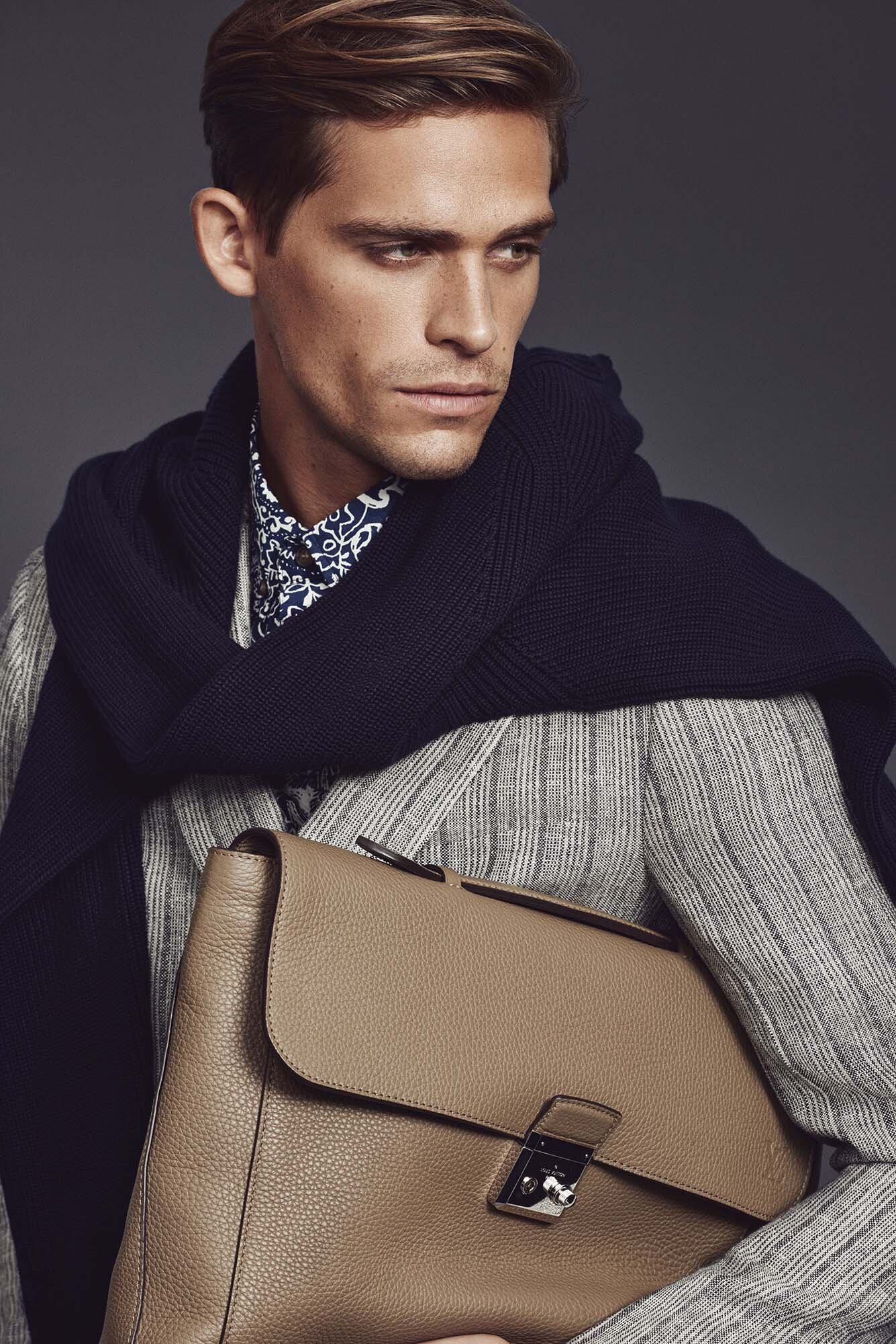 Handsome man in a patterned shirt, draped navy sweater, and striped jacket holds a tan leather briefcase looking off camera indoors.