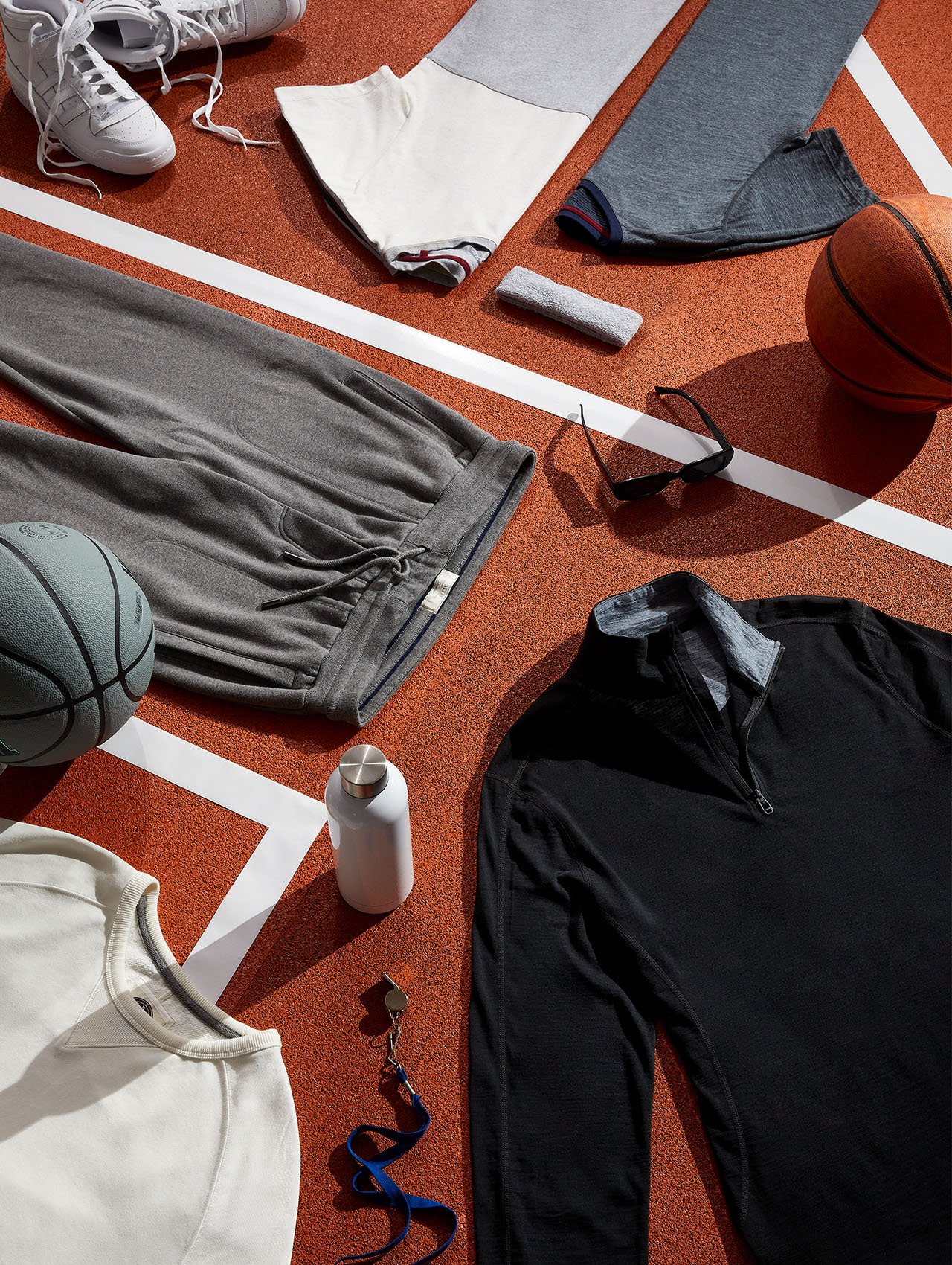 Flat lay of athletic apparel and accessories on a red running track: sneakers, workout clothes, basketball, and water bottle.