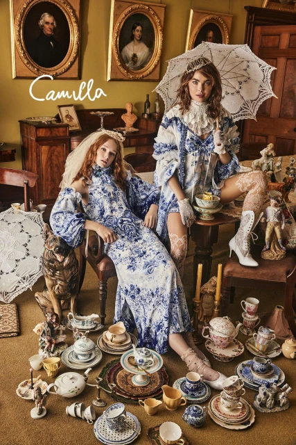 Two models in vintage-inspired fashion pose with dog amidst tea set on the floor, portraits hanging on the wall in an indoor setting.