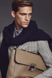 Close-up portrait of a man holding a tan leather briefcase, wearing a striped blazer and dark navy sweater.