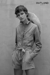 Black and white portrait of a woman with short hair wearing a button-up denim shirt and belted shorts against a textured wall.
