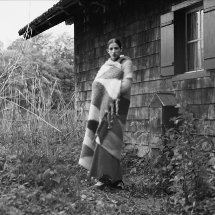 Monochrome outdoor portrait of a young woman wrapped in a striped blanket, standing near a wood-sided house in a grassy, wooded area.