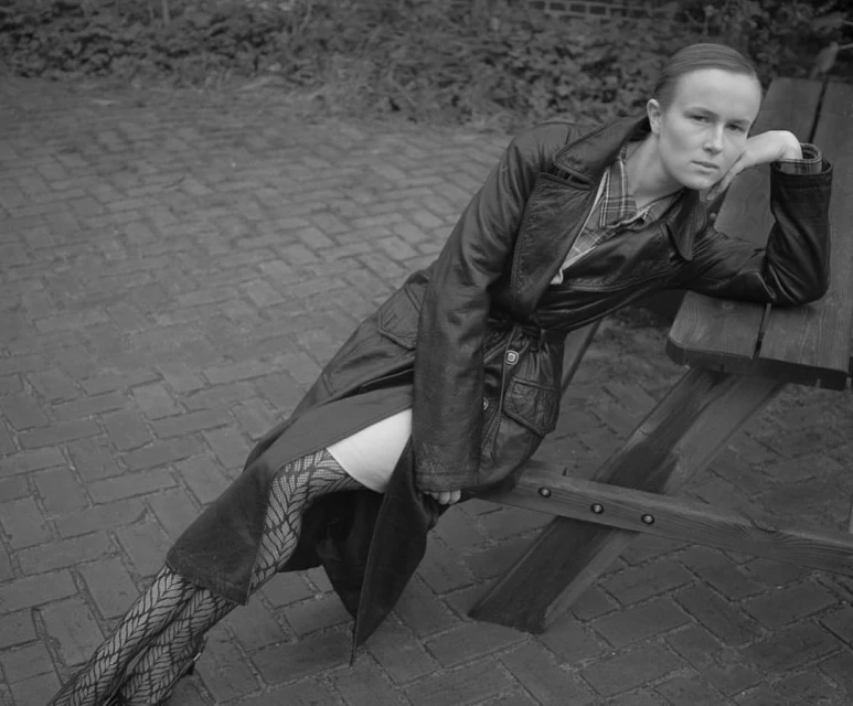 Monochrome portrait of a woman in a leather trench coat posing on a park bench in an outdoor setting with brick ground.