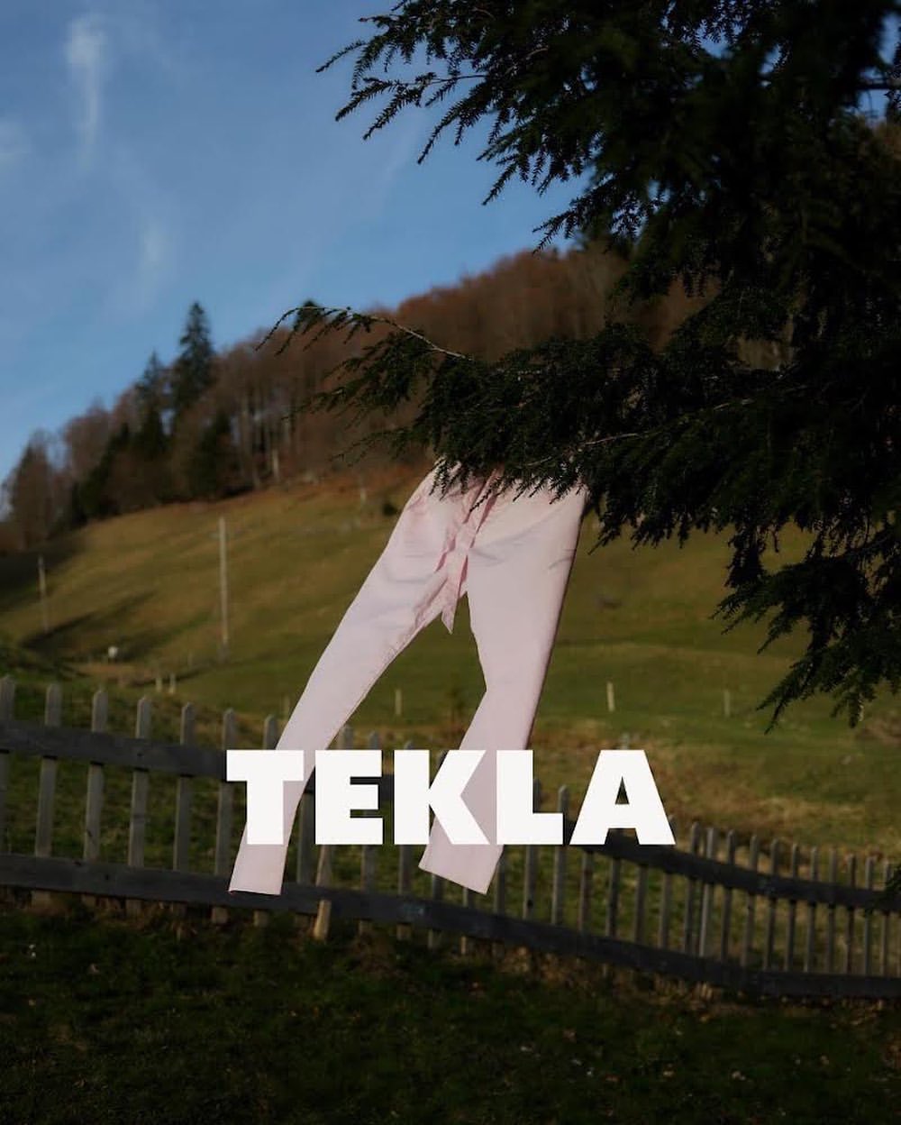 Light pink pants hang from a tree branch in front of a grassy hillside and a weathered wooden fence, with the brand name TEKLA in white.