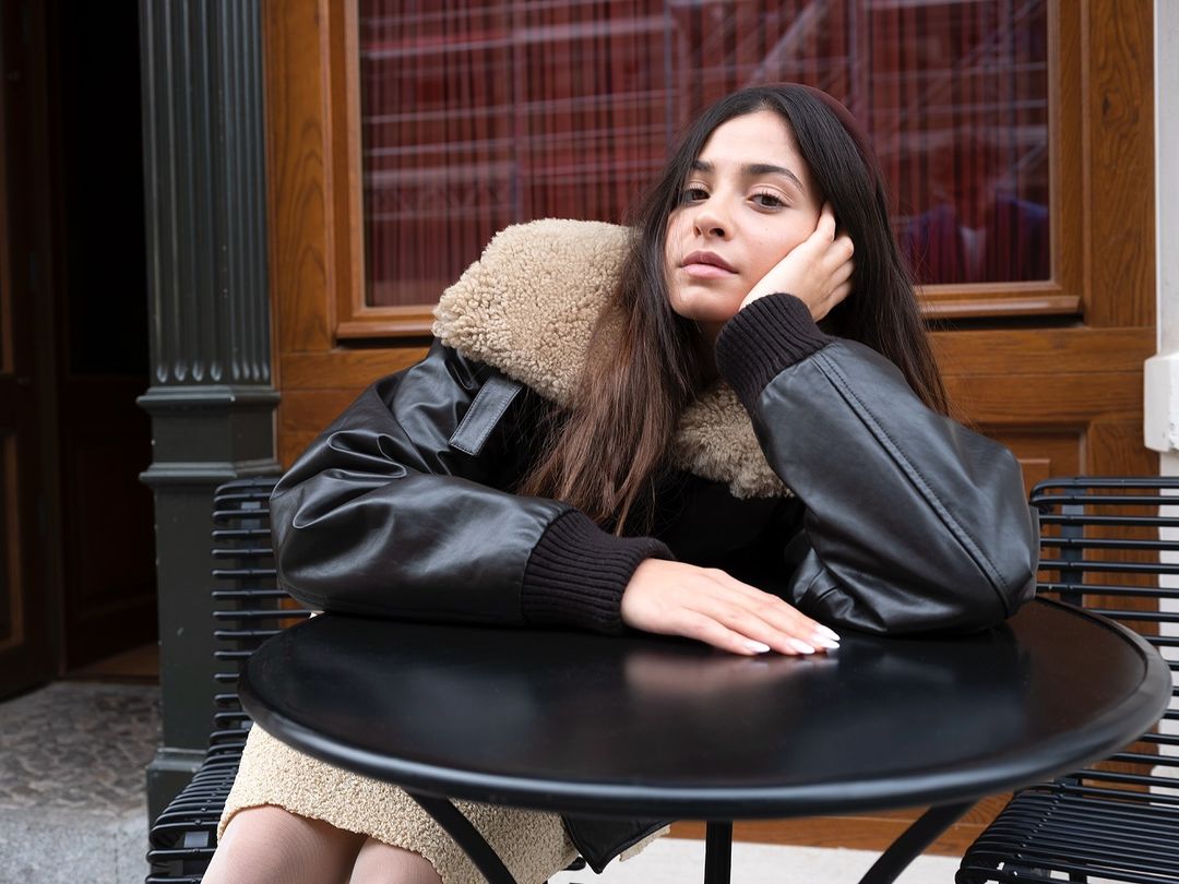 A young woman sits at an outdoor table wearing a black jacket with a fur collar, gazing into the camera in a medium shot.