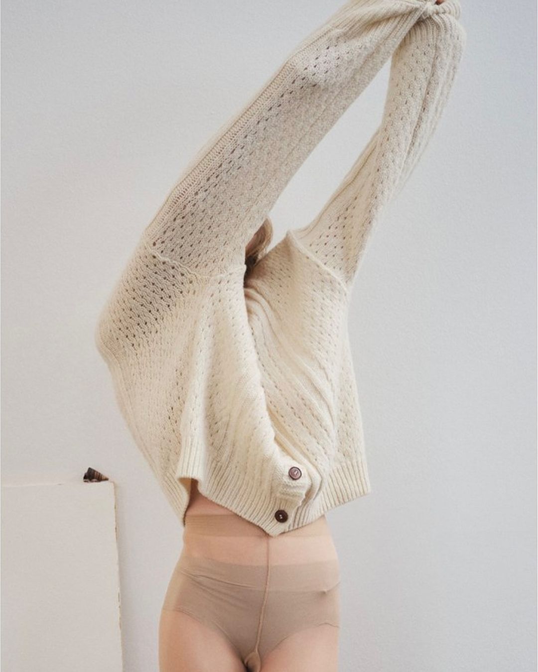Woman wearing a cream-colored, knitted cardigan and pantyhose poses with arms raised in front of a neutral-toned background.