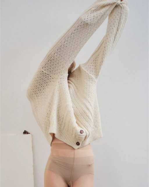 Woman wearing a cream-colored, knitted cardigan and pantyhose poses with arms raised in front of a neutral-toned background.