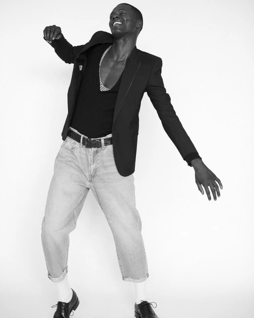 Monochrome studio shot of a Black male model in a blazer, t-shirt, cuffed jeans, socks, and dress shoes dancing against a white backdrop.