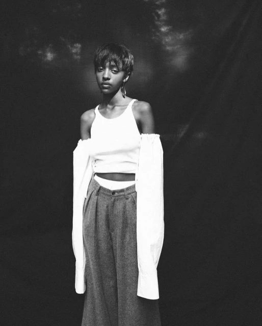 Monochrome portrait of a Black woman in a white tank top and grey slacks, against a dark backdrop, looking at the camera.