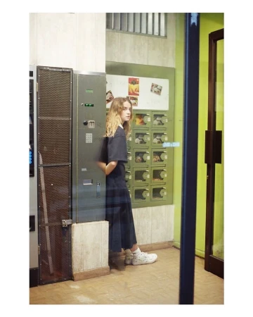 Young woman in casual dark clothing stands in an indoor setting near vending machines, looking over her shoulder with a neutral expression.