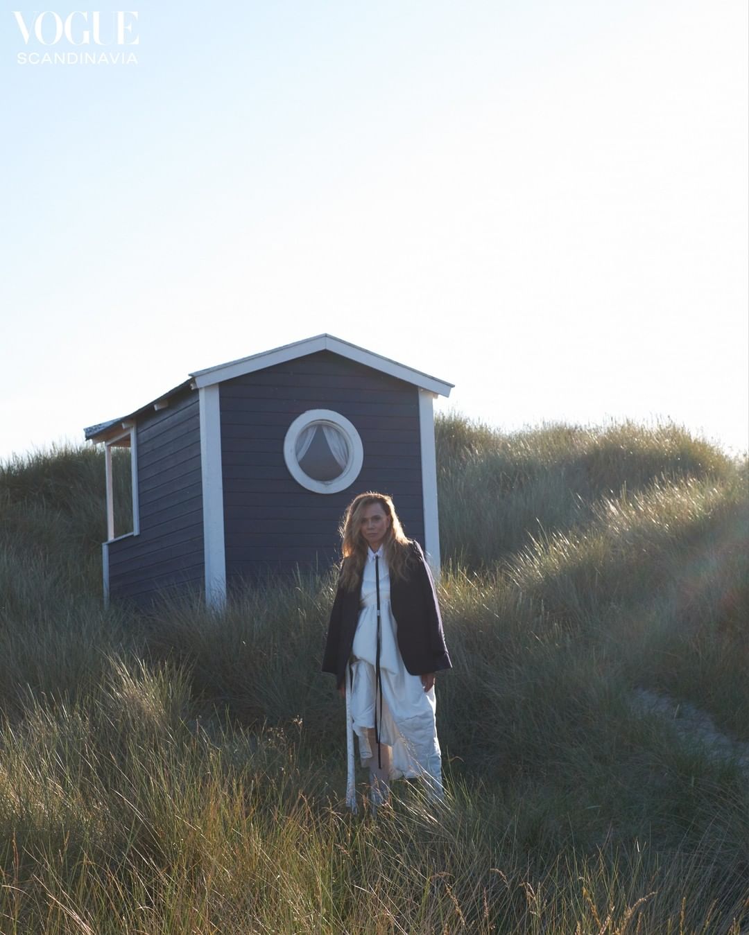 A blonde woman wearing a dark coat and white dress poses outdoors near a blue beach house with tall grass on a sunny day.