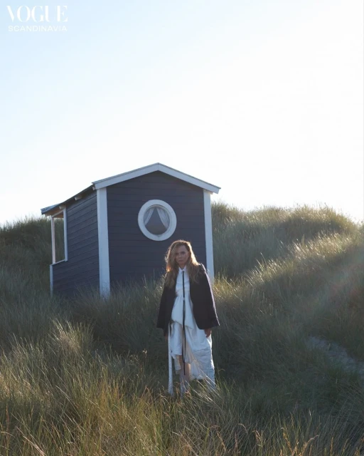 A blonde woman wearing a dark coat and white dress poses outdoors near a blue beach house with tall grass on a sunny day.
