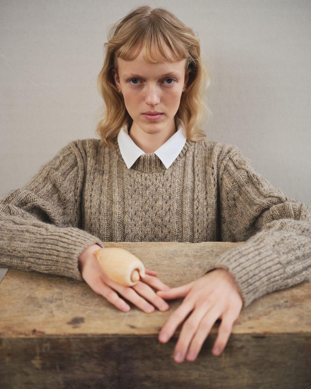 Portrait of a young woman at a wooden table, wearing a knit sweater and white collar shirt, holding a wooden object in her hands.