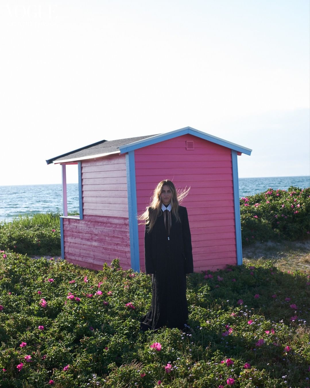 Woman in black dress standing near pink beach hut on a sunny day with ocean view, surrounded by green plants with pink flowers.
