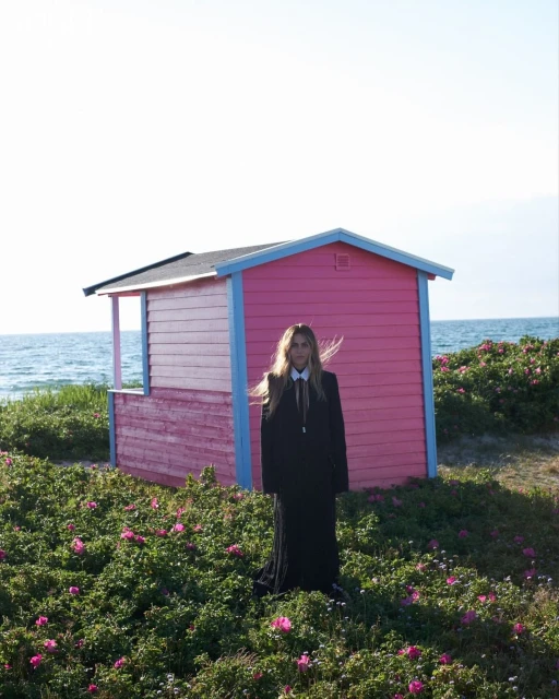 Woman in black dress standing near pink beach hut on a sunny day with ocean view, surrounded by green plants with pink flowers.