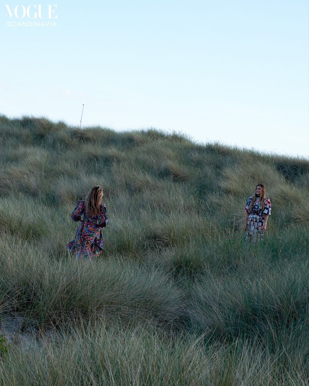 Two women in floral dresses stand in tall green grass under a light blue sky in an outdoor setting, fashion editorial, Vogue.