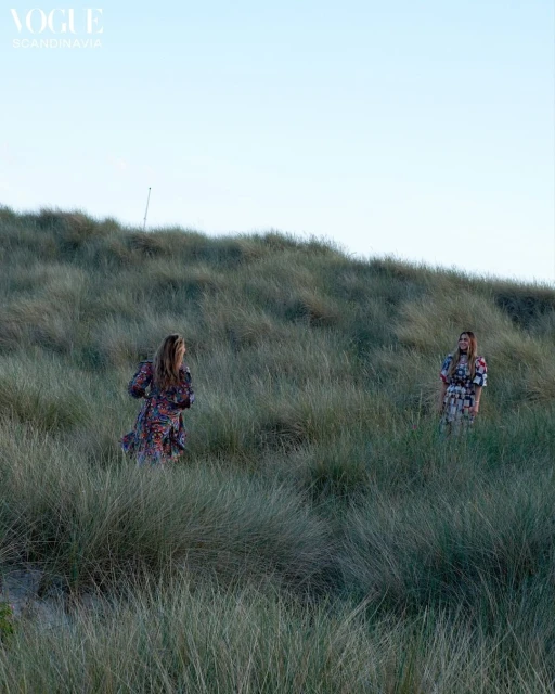 Two women in floral dresses stand in tall green grass under a light blue sky in an outdoor setting, fashion editorial, Vogue.