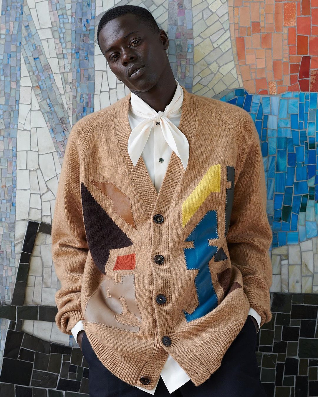 A man in a tan cardigan poses against a mosaic tile wall; his style blends casual comfort with a touch of retro sophistication.
