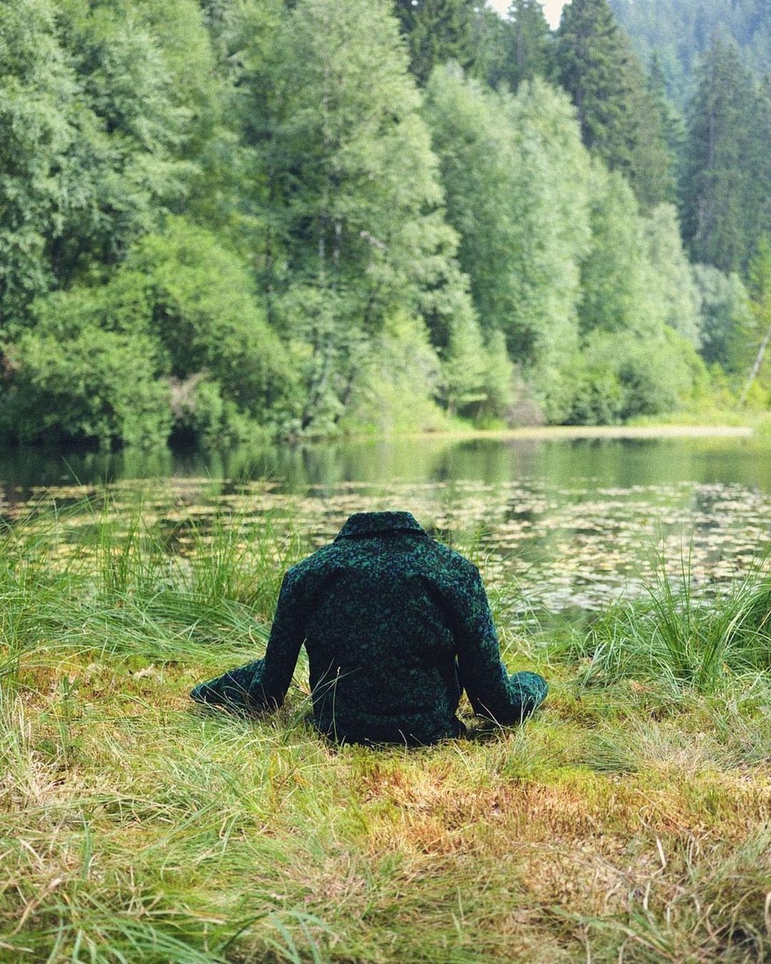 A dark green coat sits on green grass in front of a lake surrounded by lush green trees on an overcast day.