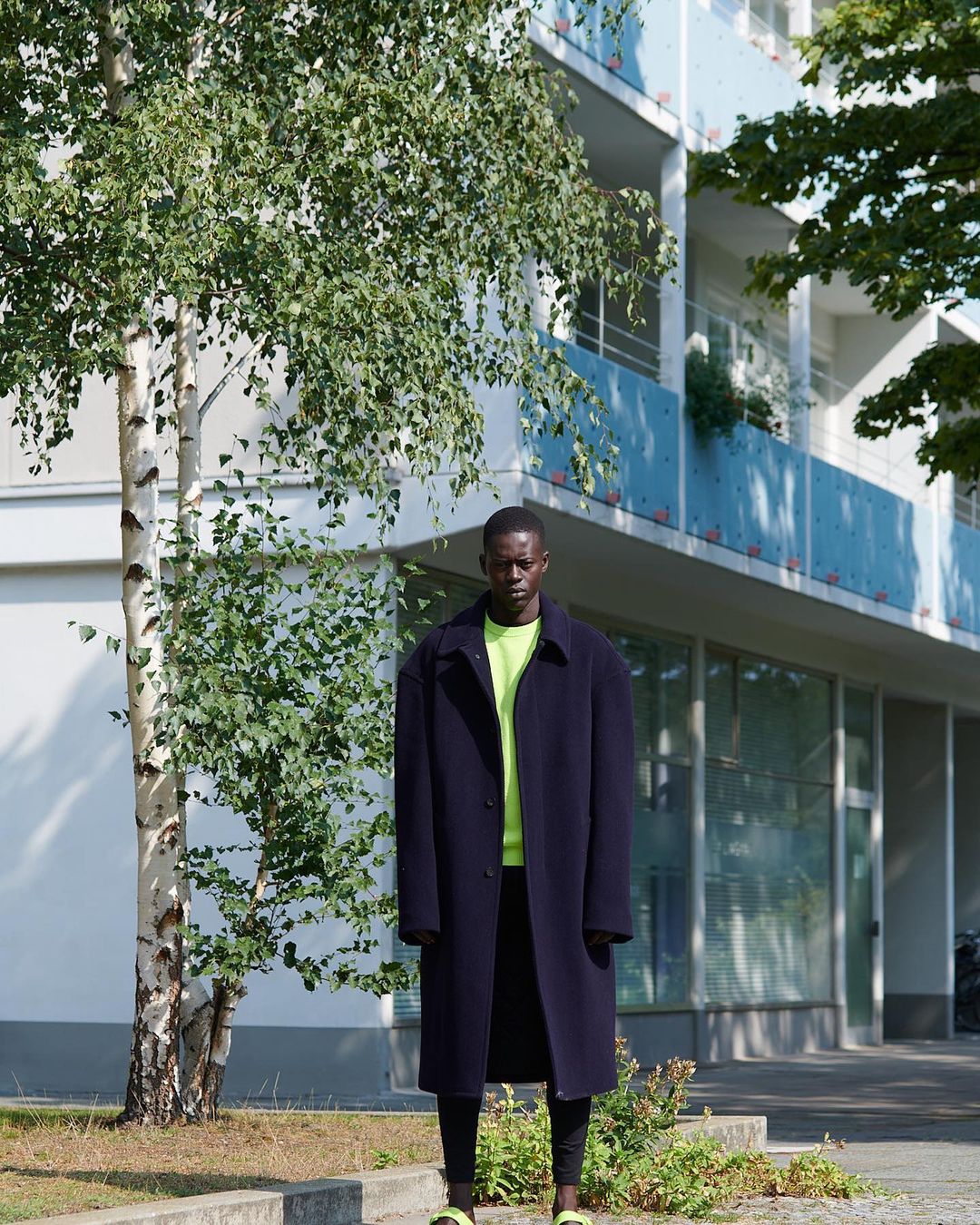 Fashion shot of a Black male model in a long dark coat and neon-yellow shirt posing outdoors by a tree and a light-blue building.