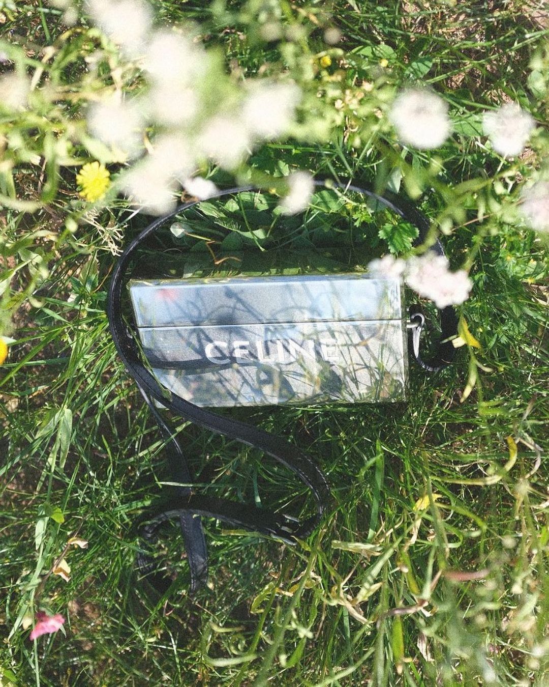 Reflective rectangular handbag resting in tall green grass with blurred white wildflowers in the foreground