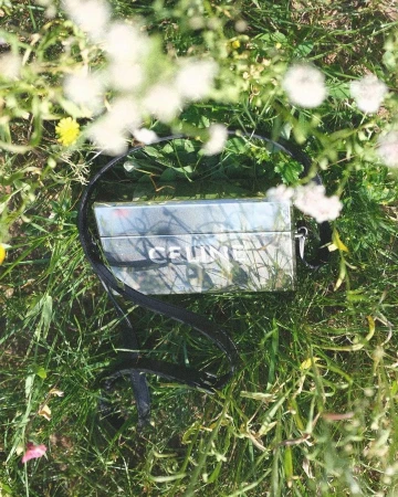Reflective rectangular handbag resting in tall green grass with blurred white wildflowers in the foreground